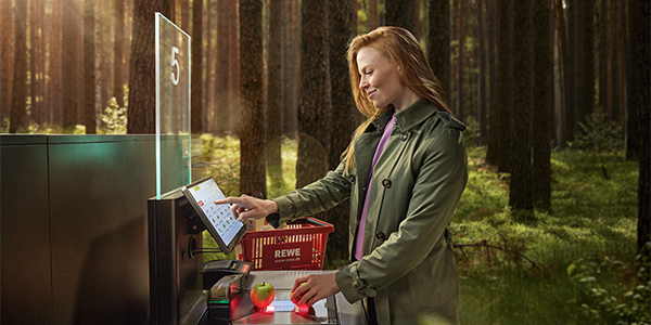 Eine moderne Selbstbedienungskasse steht mitten im Wald. Eine Person nimmt Produkte aus einem roten REWE Einkaufskorb und scannt sie ein.