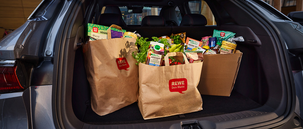 Ein Karton und zwei REWE Papiertüten gefüllt mit Lebensmitteln im Kofferraum eines Autos 