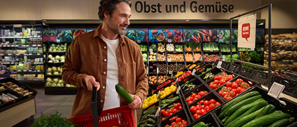 Ein lächelnder Mann im offenen Hemd füllt seinen Einkaufskorb in der REWE Gemüseabteilung 