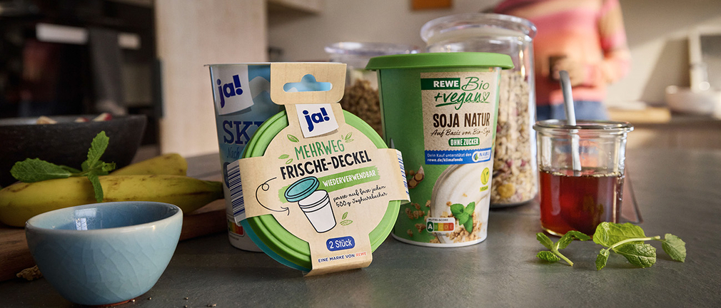 REWE Soja Naturjoghurt und Skyr, eine Schale und ein Mehrweg-Deckel auf einer Arbeitsplatte mit Bananen, Müsli und Tee   