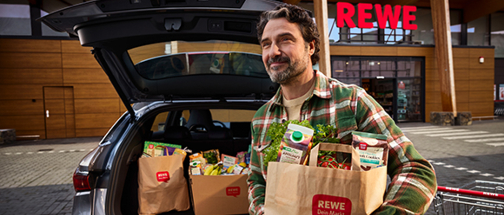 Ein Mann trägt eine mit Einkäufen gefüllte REWE Papiertüte, hinter ihm ein Kofferraum mit weiteren Einkäufen und ein REWE Markt 