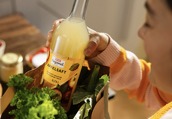 Eine Frau holt eine Flasche REWE Regional Apfelsaft auf einer REWE Papiertragetasche voll frischem Obst und Gemüse. 