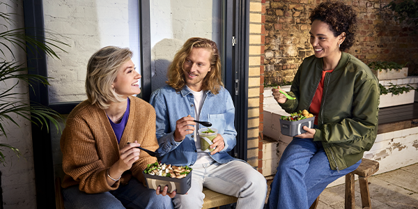 Drei Personen sitzen draußen auf einer Bank. Sie essen Salate und Suppen to go aus Mehrweg-Pfandbehältern. 