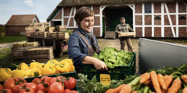 Zwei Landwirt:innen bereiten Kisten mit verschiedenen Obst- und Gemüsesorten für den Transport zu REWE vor. In den Kisten stecken gelbe Schilder mit der Aufschrift „Aus deiner Region“.