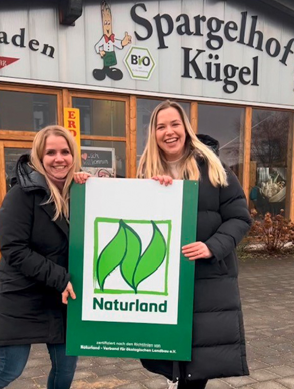 Zwei der Töchter der Familie Kügel halten zwischen sich ein großes Schild mit dem Naturland Logo. Als REWE Wegbereiter haben sie sich im Öko-Verband angemeldet. 