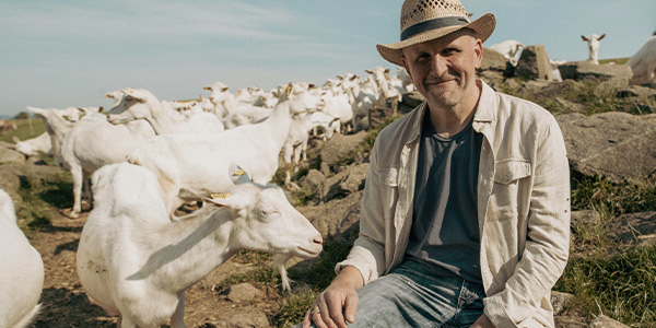 Frank Haslehner sitzt auf einem Felsen und beobachtet seine Ziegenherde. Die Tiere grasen auf einer Bergwiese. 