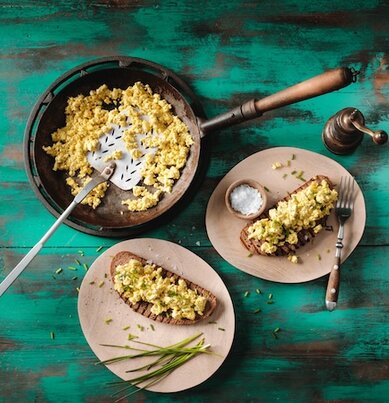 Eine Pfanne mit veganem Rührei steht auf einem gedeckten Holztisch. 