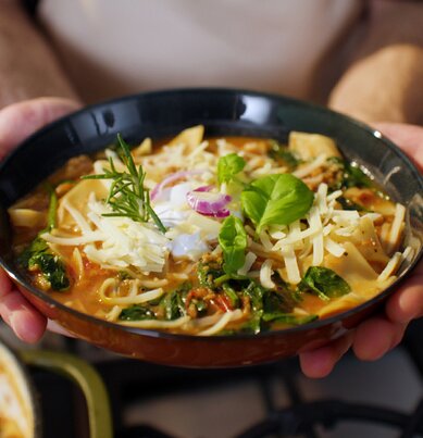 Eine Schüssel Tomaten-Suppe mit darin gekochten Lasagne-Platten, geriebenem Käse, frischen Basilikum und Rosmarin. 