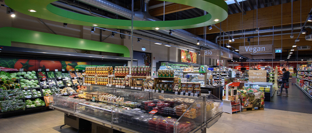 Von der Obst-und Gemüseabteilung aus schaut man in einen Green Building Markt. Im Hintergrund sieht man Regale mit vegetarischen, veganen und Bio-Produkten. 