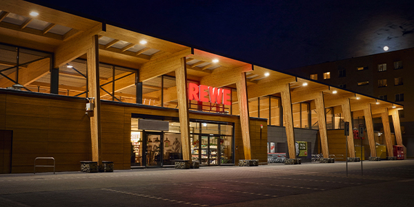 Der Eingang zu einem beleuchteten REWE Green Building in der Dämmerung. Auf dem Dach kann man Solarpanels erkennen. 