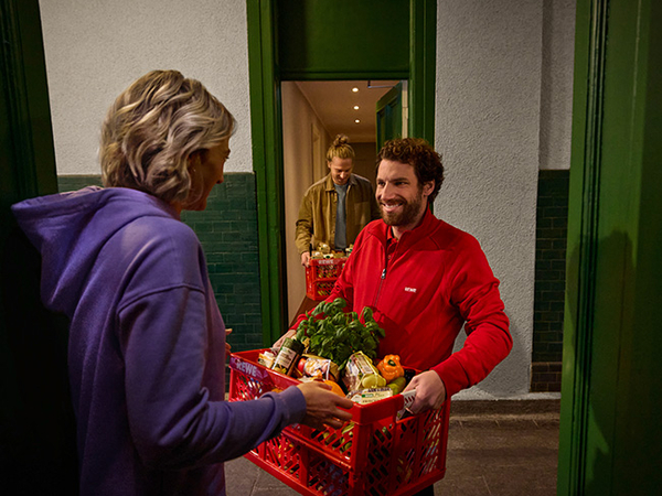 Ein Mitarbeiter des REWE Lieferservice übergibt einer Kundin ihre Bestellung an der Wohnungstür.