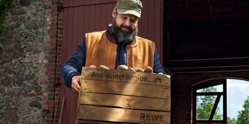 Ein bauer, der REWE beliefert, trägt eine Kiste mit Kartoffeln.
