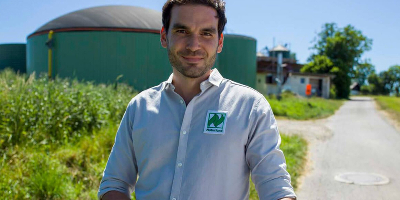 Mann mit Hemd steht draußen vor einer grünen Landwirtschaftsfläche mit Biogas-Anlage