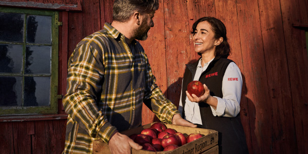 Eine REWE Mitarbeiterin im Gespräch mit einem regionalen Landwirt. Die Darstellung vermittelt: Sie weiß, woher die regionalen Äpfel in ihrem REWE Markt kommen und geht partnerschaftlich mit den Erzeuger:innen um. 