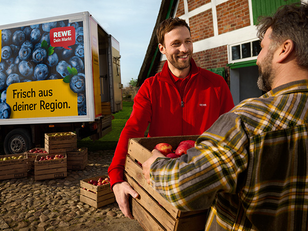 Ein Fahrzeug des REWE Lieferservice steht auf einem regionalen Hof. Auf der Seite ist der Schriftzug „Aus deiner Region“ zu erkennen. Der Landwirt gibt dem REWE Lieferservice Mitarbeiter eine Kiste mit frischen Äpfeln. 