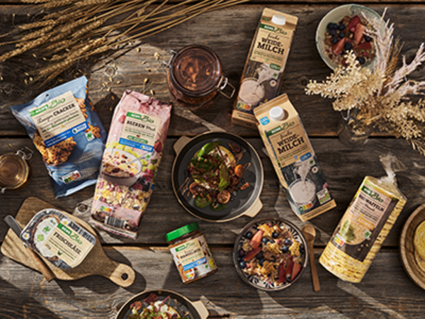 Eine Auswahl an REWE Bio Produkten mit Naturland Siegel liegen auf einem rustikalen Holztisch. Dazwischen stehen mehrere Schüsseln mit unterschiedlichen Gerichten und reife Kornähren.