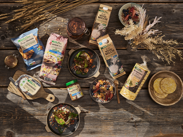 Eine Auswahl an REWE Bio Produkten mit Naturland Siegel liegen auf einem rustikalen Holztisch. Dazwischen stehen mehrere Schüsseln mit unterschiedlichen Gerichten und reife Kornähren.