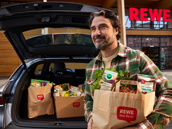 Auf dem Parkplatz vor einem REWE Markt: Eine Person lädt volle Einkaufstüten in den Kofferraum. 