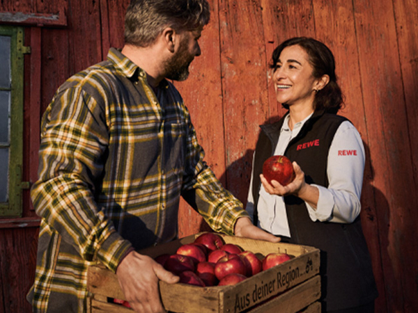 Eine REWE Mitarbeiterin im Gespräch mit einem regionalen Landwirt. Die Darstellung vermittelt: Sie weiß, woher die regionalen Äpfel in ihrem REWE Markt kommen und geht partnerschaftlich mit den Erzeuger:innen um. 
