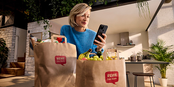 Eine Person steht mit vollen REWE Einkaufstüten in der Küche. Sie schaut aufs Handy.