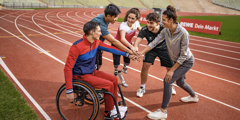 Menschen mit und ohne Behinderung legen ihre Hände aufeinander, um sich auf einer Tartanbahn auf ein sportliches Event einzustimmen. 