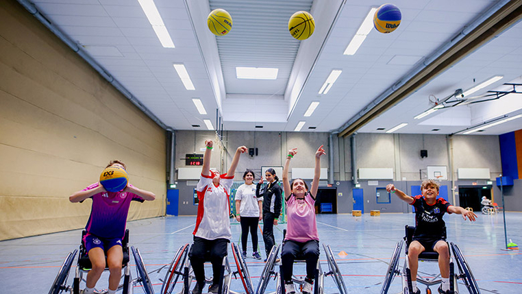 Mehrere Kinder im Rollstuhl werfen auf einen Basketballkorb. 