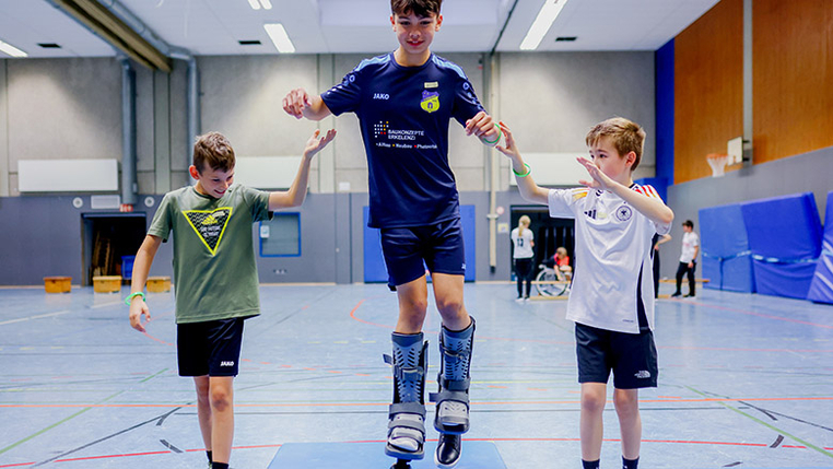 Ein Kind trägt Beinprothesen und balanciert damit auf dem Oberteil eines Turnkastens. Zwei andere Kinder helfen beim Gleichgewicht halten. 