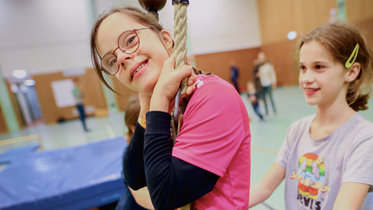 Ein Kind mit Behinderung schwingt in einer Turnhalle an einem Tau. Ein zweites Kind hilft ihm. 