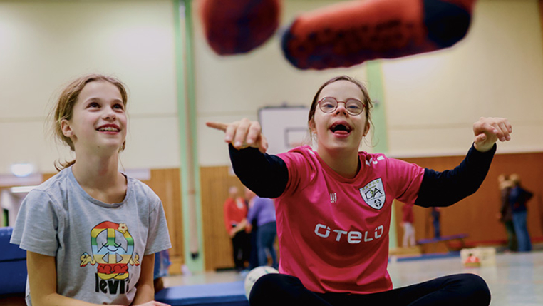 In einer Sporthalle. Zwei lachende Kinder mit und ohne Behinderung machen gemeinsam eine Übung. 