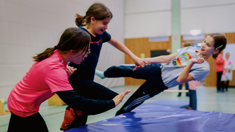In einer Sporthalle. Drei Kinder mit und ohne Behinderung lassen sich gemeinsam auf eine dicke Gymnastikmatte fallen. 