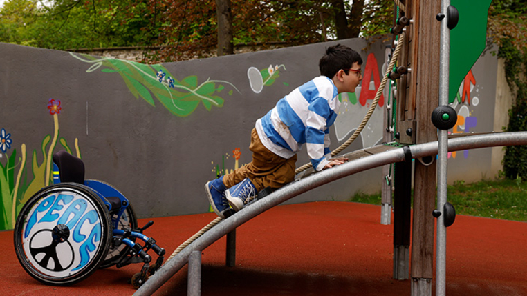 Ein Kind klettert aus dem Rollstuhl direkt auf ein inklusives Spielgerät. 