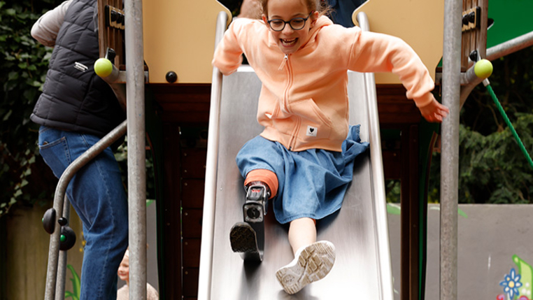 Ein Kind mit Beinprothese lacht auf der inklusiven Rutsche.  