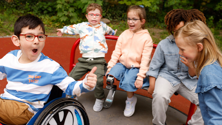 Kinder mit und ohne Behinderung auf einem inklusiven Karussell. In der Mitte sitzt ein Kind im Rollstuhl und zeigt „Daumen hoch“. 