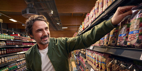 Ein Mann im Supermarkt. Sein Einkaufskorb ist gefüllt mit REWE Bio Produkten und er greift nach einer Packung REWE Bio Crunchy im Regal.  