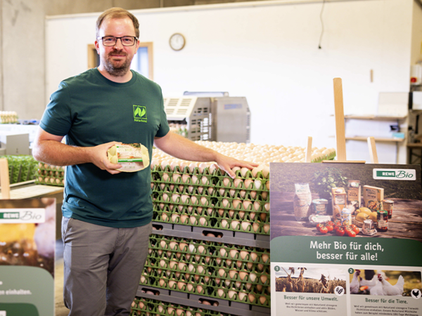 Christoph Möller hält eine Packung REWE Spitz & Bube Eier in der Hand. Ein großes Plakat von REWE Bio trägt die Aufschrift „Besser für unsere Tiere“. 