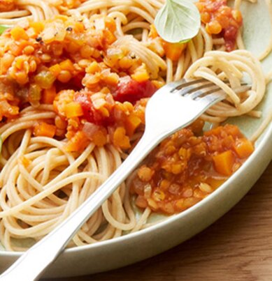 Zwei Teller mit Spaghetti und Linsenbolognese, eine Pfanne mit Linsenbolognese, zwei kleine Teller mit Salz und frischem Basilikum. In der Mitte liegt eine Packung REWE Bio Vollkorn Spaghetti. 
