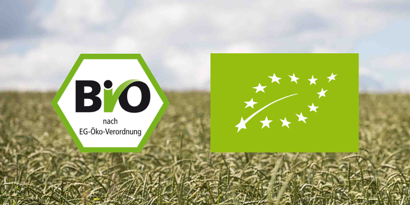 Das EU-Bio-Logo und das deutsche Bio-Siegel vor dem Hintergrund eines Getreidefeldes. 