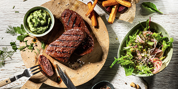 Auf einem Holzbrett stehen gegrillte pflanzliche Steaks mit einem kleinen Salat und Dipp.